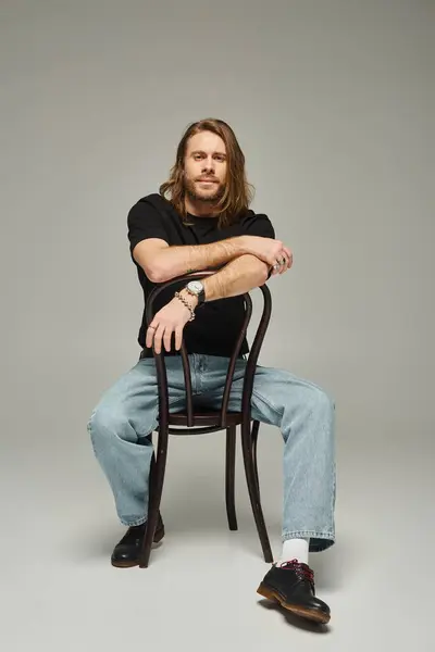 Pleine longueur de barbu bel homme avec les cheveux longs assis en jeans et t-shirt sur chaise sur gris — Photo de stock
