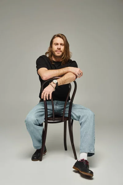 Full length of bearded good looking man with long hair sitting in jeans and t-shirt on chair on grey — Stock Photo