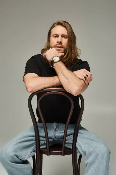Hombre barbudo y guapo con el pelo largo sentado en jeans y camiseta en silla de madera en gris - foto de stock