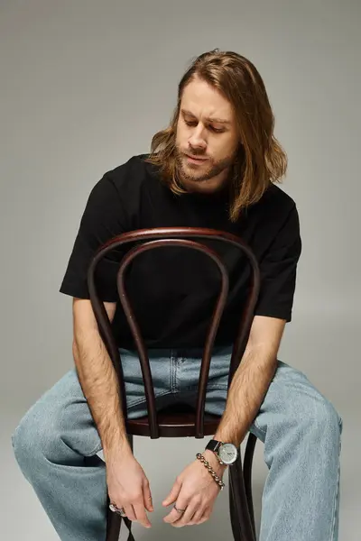 Bearded good looking man with long hair sitting in jeans and t-shirt on wooden chair on grey — Stock Photo