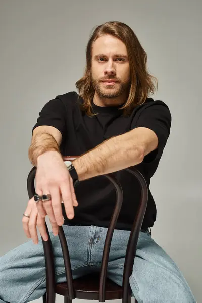Бородатий і красивий чоловік з довгим волоссям, сидячи в джинсах і футболці на стільці на сірому фоні — стокове фото