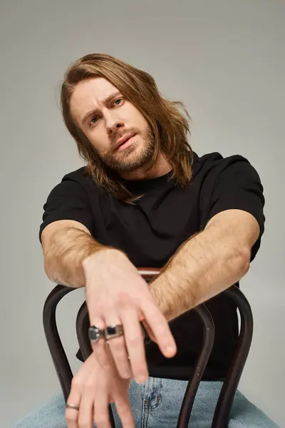 Bärtiger, gutaussehender Mann mit langen Haaren sitzt in Jeans und T-Shirt auf einem Holzstuhl in grau — Stockfoto