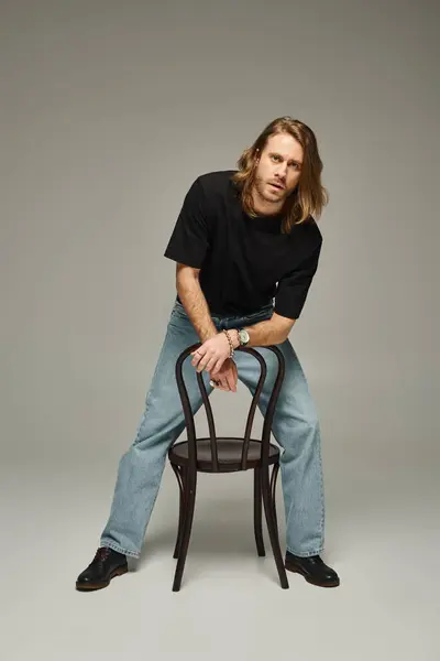Volle Länge des bärtigen gut aussehenden Mannes mit langen Haaren in Jeans und T-Shirt sitzt auf Stuhl auf grau — Stockfoto