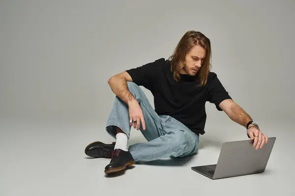 Handsome man with long hair and beard sitting in denim jeans on floor and using laptop on grey — Stock Photo