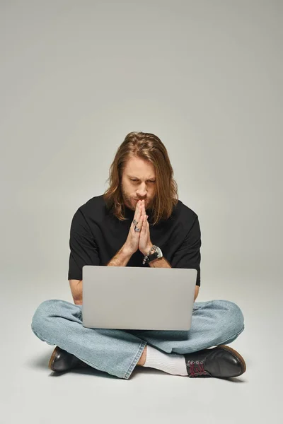 Handsome man with long hair and beard sitting with crossed legs on floor and using laptop on grey — Stock Photo