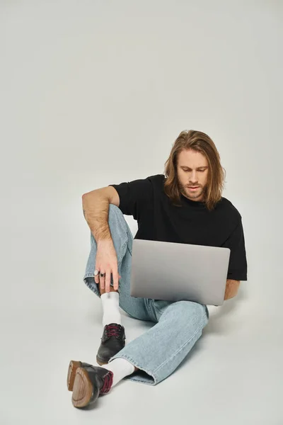 Bel freelance con capelli lunghi e barba seduta sul pavimento e con computer portatile su sfondo grigio — Foto stock
