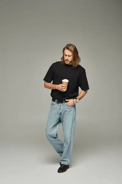 Hombre guapo con pelo largo y barba sosteniendo taza de papel con café para ir sobre fondo gris - foto de stock