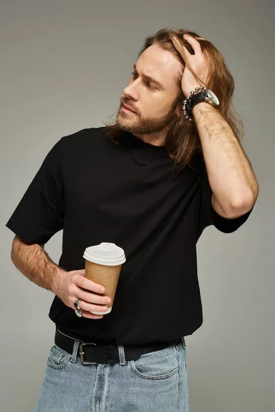 Красивий чоловік з довгим волоссям і бородою тримає паперову чашку з кавою, щоб піти на сірий фон — стокове фото