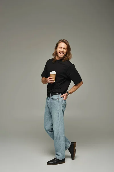 Homme gai avec les cheveux longs et la barbe tenant tasse en papier avec café pour aller sur fond gris — Photo de stock