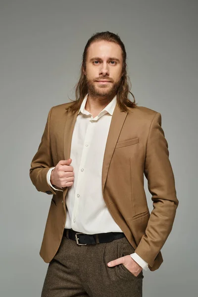 Uomo d'affari con barba e capelli lunghi in posa con mano in tasca su sfondo grigio, abbigliamento formale — Foto stock