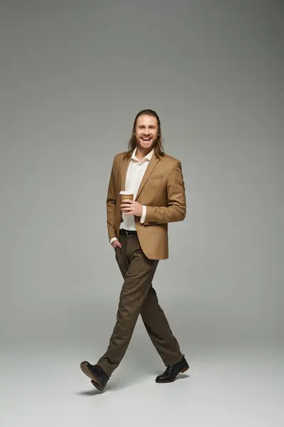 Homme d'affaires heureux avec barbe et cheveux longs tenant café pour aller sur fond gris, tenue formelle — Photo de stock