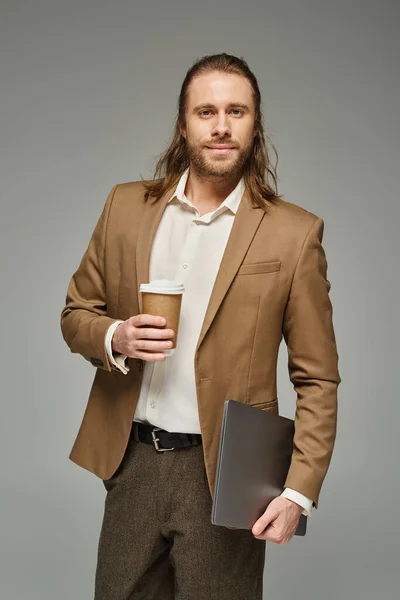 Красивый бизнесмен с бородой и длинными волосами держит кофе, чтобы пойти и ноутбук на сером фоне — стоковое фото