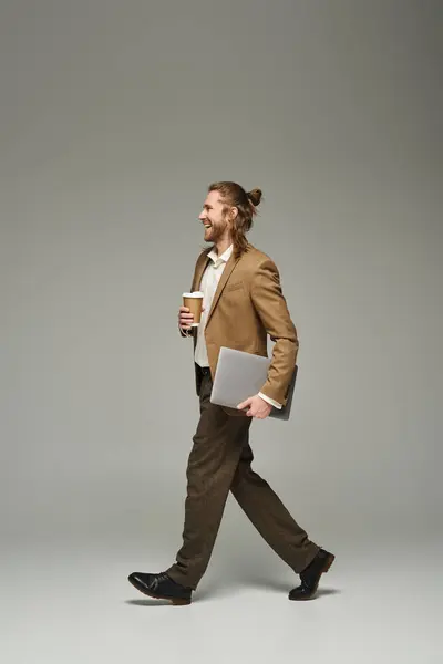 Uomo d'affari felice con barba e capelli lunghi che tengono il caffè per andare e laptop mentre cammina sul grigio — Foto stock