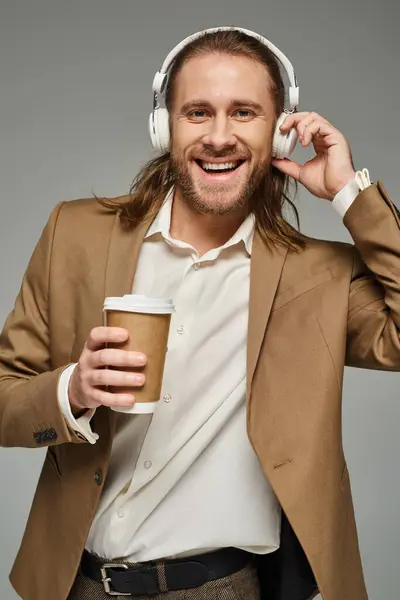 Happy bearded businessman in headphones holding coffee to go on grey backdrop, formal attire — Stock Photo