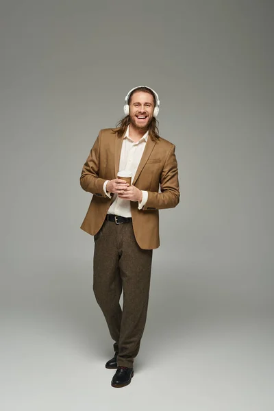 Allegro uomo d'affari barbuto in cuffia che tiene il caffè per andare su sfondo grigio, abbigliamento formale — Foto stock