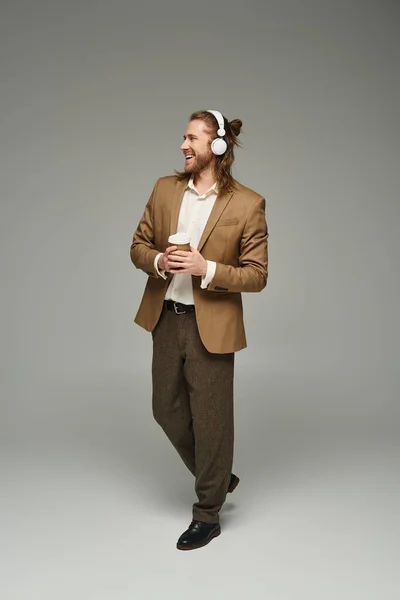 Empresário barbudo alegre em fones de ouvido segurando café para ir em pano de fundo cinza, traje formal — Fotografia de Stock