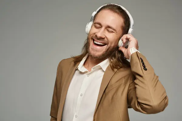 Изумленный бородатый бизнесмен в наушниках слушает музыку на сером фоне, формальный наряд — стоковое фото