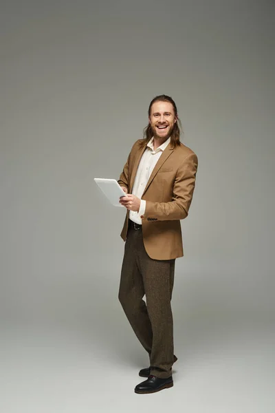 Happy businessman with long hair posing in formal attire and using digital tablet on grey backdrop — Stock Photo