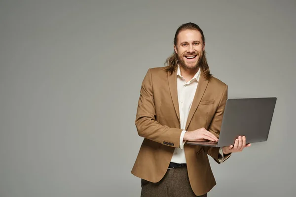 Homem de negócios feliz com cabelos longos posando em traje formal e usando laptop em pano de fundo cinza — Fotografia de Stock
