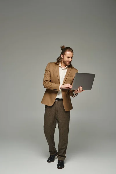 Handsome businessman with long hair posing in formal attire and using laptop on grey backdrop — Stock Photo