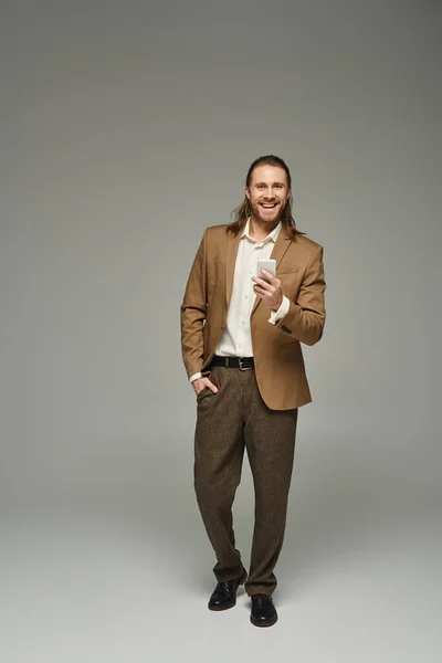 Bonito e feliz barbudo empresário em traje formal usando smartphone no fundo cinza — Fotografia de Stock
