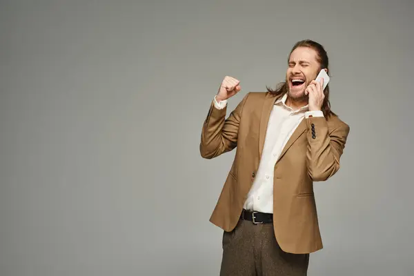 Handsome and excited bearded businessman in formal attire talking on smartphone on grey backdrop — Stock Photo