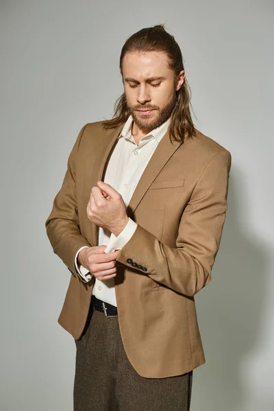 Hombre de negocios guapo con el pelo largo y la barba ajustando la manga en blazer beige sobre fondo gris - foto de stock