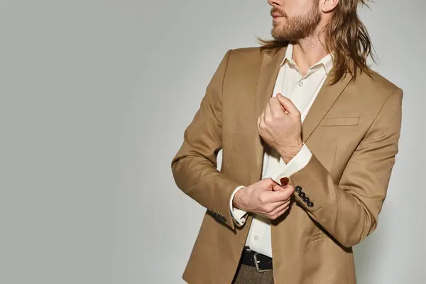 Good looking businessman with long hair and beard adjusting sleeve on beige blazer on grey backdrop — Stock Photo