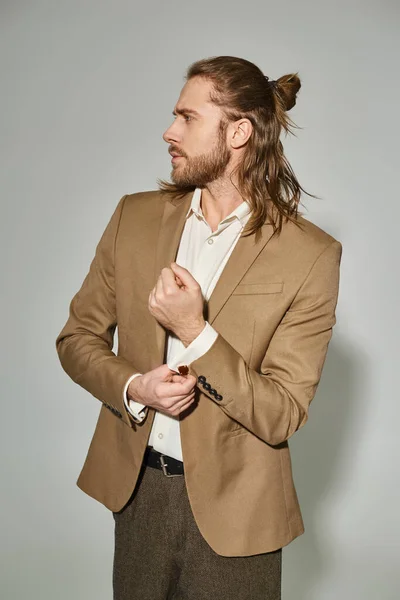 Homem de negócios de boa aparência com cabelos longos e barba ajustando a manga no blazer bege no fundo cinza — Fotografia de Stock