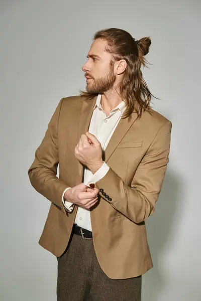 Good looking businessman with long hair and beard adjusting sleeve on beige blazer on grey backdrop — Stock Photo