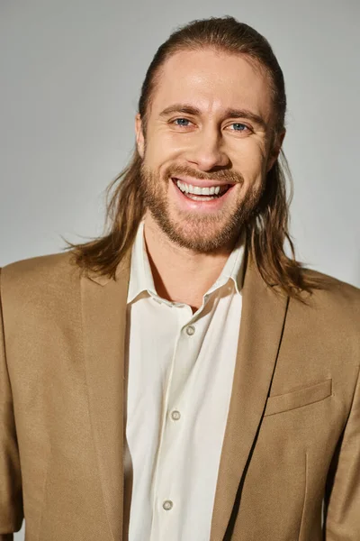 Portrait of positive businessman with long hair and beard posing in beige blazer on grey backdrop — Stock Photo