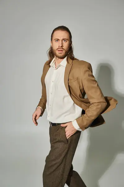 Schöner Geschäftsmann mit langen Haaren und Bart posiert mit der Hand in der Tasche auf grauem Hintergrund — Stockfoto