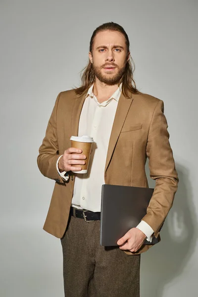 Gutaussehender und bärtiger Geschäftsmann in festlicher Kleidung mit Laptop und Kaffee vor grauem Hintergrund — Stockfoto