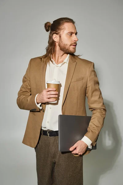 Bonito e barbudo empresário em traje formal segurando laptop e café no fundo cinza — Fotografia de Stock