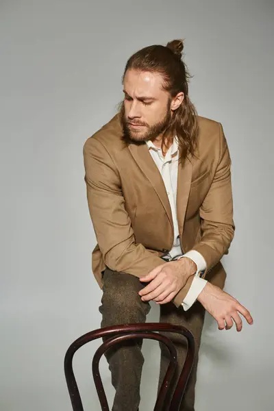 Atraente barbudo empresário em bege traje formal posando perto de cadeira de madeira no fundo cinza — Fotografia de Stock