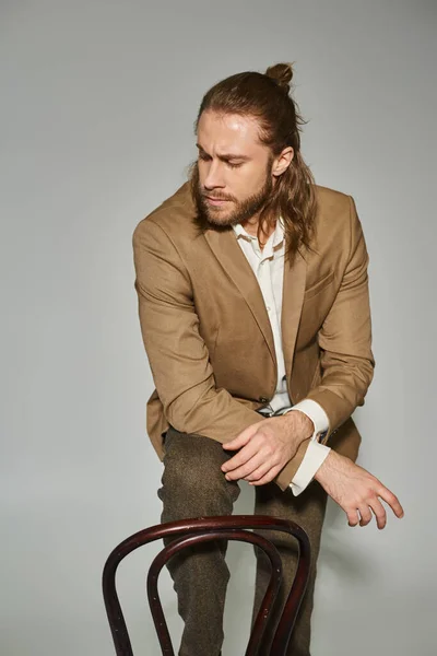 Attractive bearded businessman in beige formal attire posing near wooden chair on grey background — Stock Photo