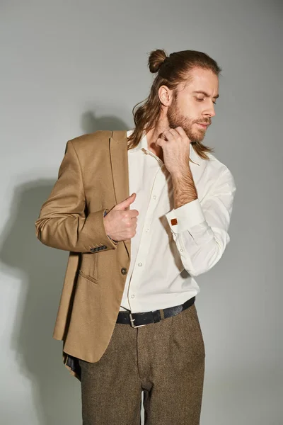 Homem de negócios barbudo de boa aparência em camisa branca posando com blazer bege no ombro no cinza — Fotografia de Stock
