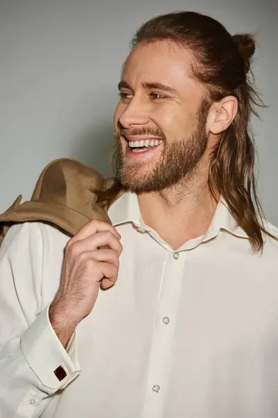 Fröhliche und bärtige Geschäftsmann in weißem Hemd posiert mit beigem Blazer auf Schulter auf grau — Stockfoto