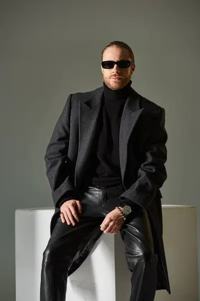 Bello uomo barbuto in occhiali da sole alla moda e cappotto di lana posa vicino cubo bianco su sfondo grigio — Foto stock