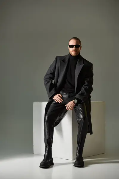 Bell'uomo barbuto in eleganti occhiali da sole e cappotto di lana seduto su cubo bianco su sfondo grigio — Foto stock