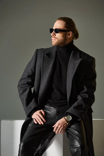 Handsome bearded man in stylish sunglasses and woolen coat sitting on white cube on grey backdrop — Stock Photo