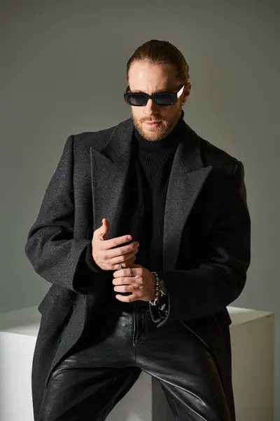 Good looking bearded man in stylish sunglasses and coat sitting on white cube on grey backdrop — Stock Photo