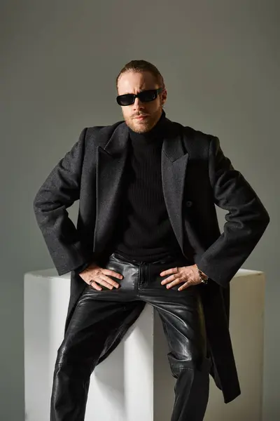 Bell'uomo barbuto in occhiali da sole alla moda e cappotto seduto su cubo bianco su sfondo grigio — Foto stock