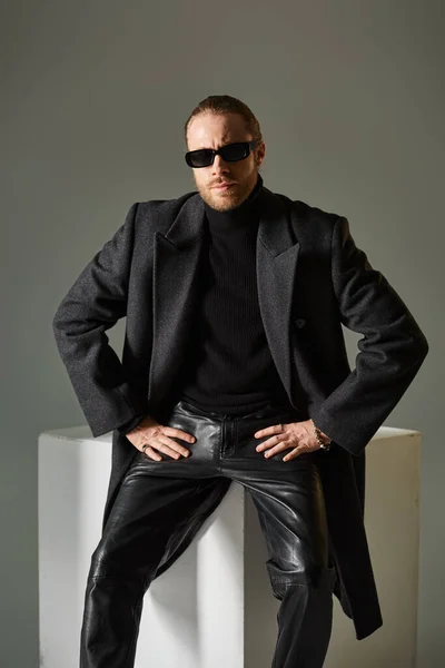 Good looking bearded man in stylish sunglasses and coat sitting on white cube on grey backdrop — Stock Photo