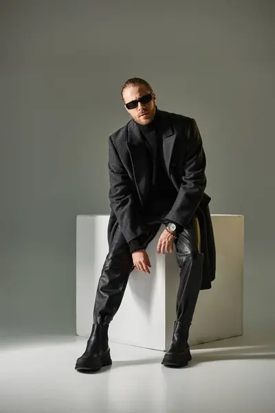 Full length of bearded man in stylish sunglasses and coat sitting on white cube on grey backdrop — Stock Photo