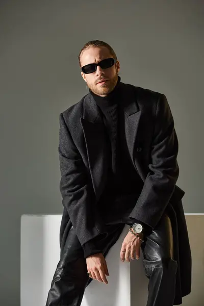 Bell'uomo elegante in occhiali da sole alla moda e cappotto seduto su cubo bianco su sfondo grigio — Foto stock