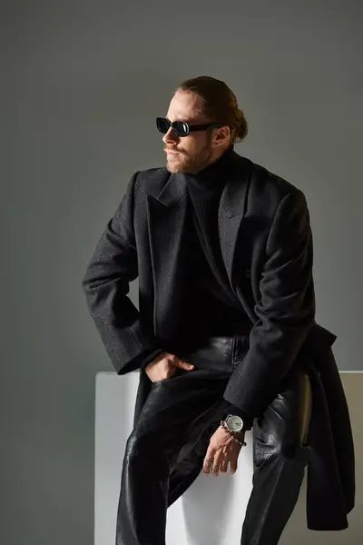 Good looking stylish man in trendy sunglasses and coat sitting on white cube on grey backdrop — Stock Photo