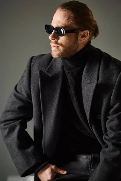 Portrait of good looking stylish man in sunglasses and coat sitting on white cube on grey backdrop — Stock Photo