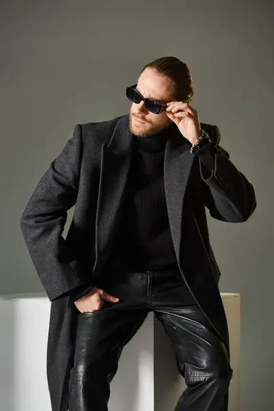 Portrait of fashionable man in trendy sunglasses and coat sitting on white cube on grey backdrop — Stock Photo