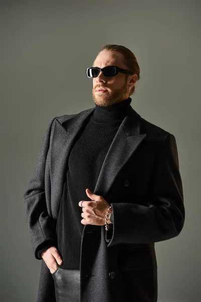 Portrait of fashionable man in trendy sunglasses and coat posing with hand in pocket on grey — Stock Photo